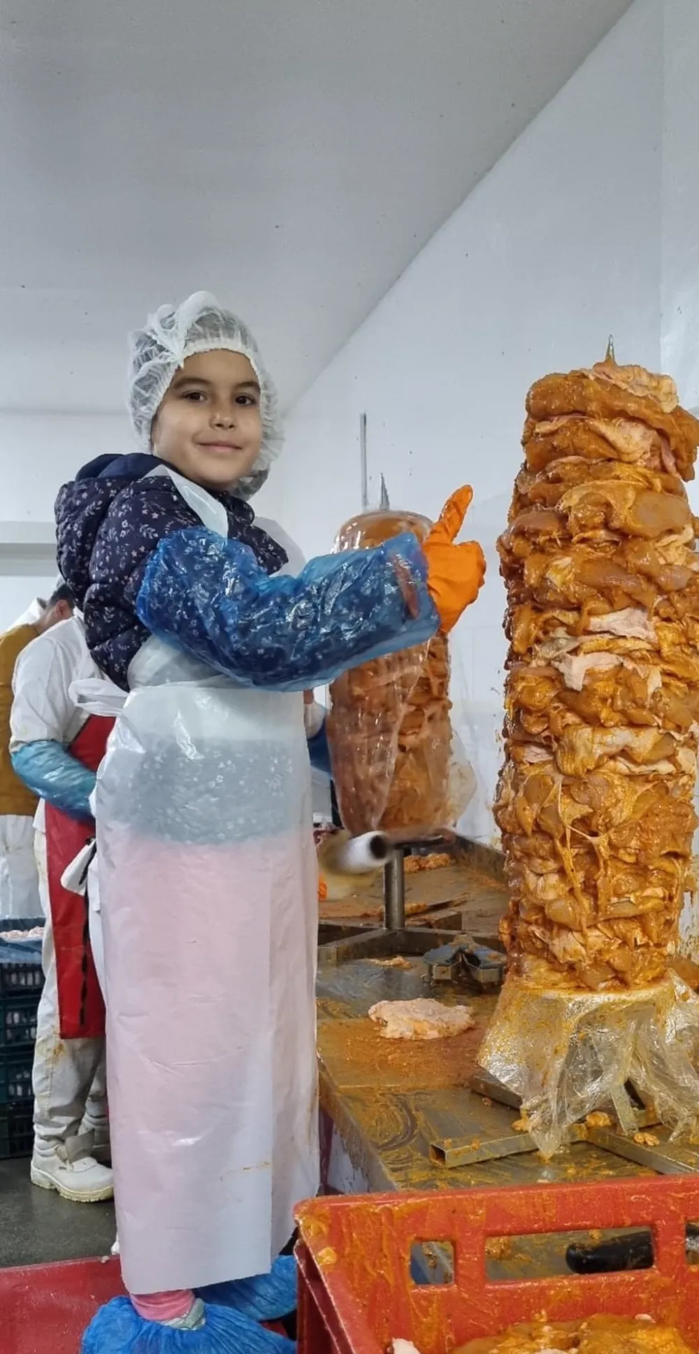 Üretim salonunda bone ve koruyucu eldivenli küçük bir kız, bir dönerin yanında başparmağını kaldırıyor.